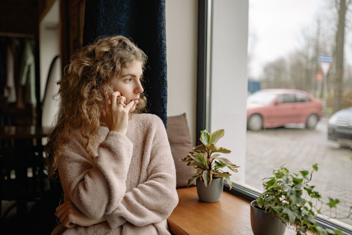Frau blickt nachdenklich aus dem Fenster, Smartphone in der Hand